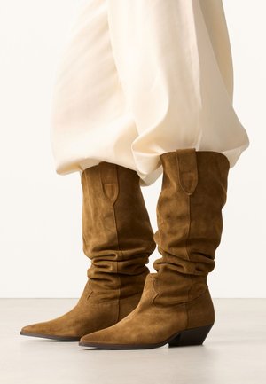 Bottes souples en daim marron mi-mollet avec bouts pointus et talons bas inclinés, portées avec un pantalon ample de couleur crème.