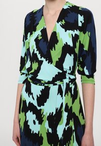 Šaty ve stylu wrap s abstraktním vzorem v modré, zelené a černé barvě. Vyrobené z elastického materiálu s hlubokým výstřihem do V a třičtvrtinovými rukávy. Detaily vázaného pasu.