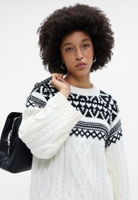 Valge kaeluseline kampsun musta mustriga õlavarrega, millel on geomeetrilised mustrid ja pikad varrukad. Kaasas must kott.