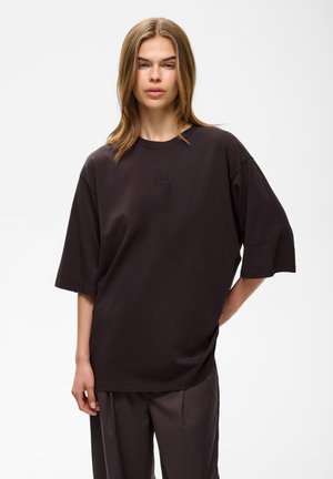 Jeune personne aux cheveux longs et raides porte une chemise courte sombre surdimensionnée et un pantalon ample assorti, debout avec une main sur la hanche.