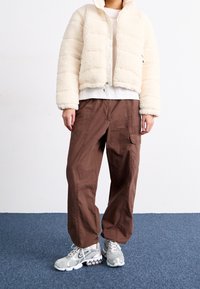 Biała, teksturowana kurtka polarowa z pionowymi szwami, brązowe spodnie cargo i szare sneakersy z czarnymi akcentami na niebieskim tle wykładziny.