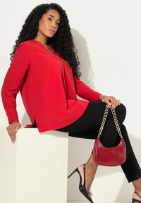 Maglietta rossa a maniche lunghe con orlo leggermente asimmetrico, abbinata a pantaloni neri slim. Presenta una borsa bordeaux con stampa a coccodrillo e tracolla in oro.