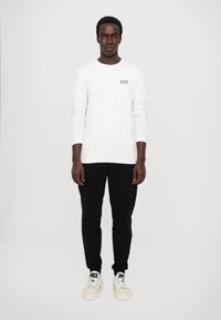 Langermet hvit skjorte med EA7-logo, kombinert med svarte joggebukser og hvite joggesko. Enkel, moderne design med en avslappet passform.