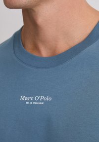 Niebieska bawełniana koszulka z okrągłym dekoltem, krótkimi rękawami oraz białym napisem z logo „Marc O’Polo EST. IN STOCKHOLM” na przodzie.