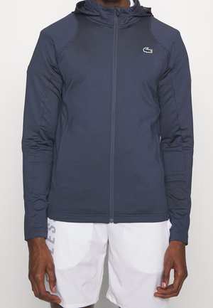 Mężczyzna w granatowej bluzie z kapturem na zamek i białych szortach, stojący na jednolitym tle.