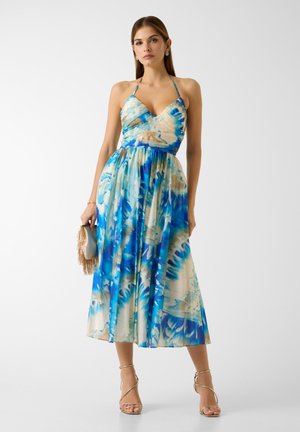 Femme portant une robe midi halter tie-dye bleue et beige et des talons dorés à brides fines, tenant une pochette beige à franges sur fond blanc.