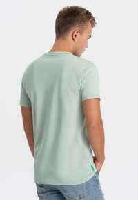 Tyrkysové tričko s krátkým rukávem vyrobené z měkké látky, s kulatým výstřihem a malou zelenou nálepkou na dolní straně.