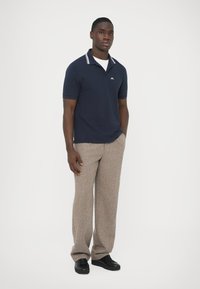 Polo bleu marine avec des rayures blanches sur le col, associé à un pantalon beige texturé et des chaussures noires, posé sur un fond blanc.