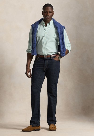 Mężczyzna stoi, ubrany w jasnosiadą pasiastą koszulę na guziki, ciemne dżinsy, brązowe mokasyny, zegarek i niebieski sweter zarzucony na ramiona.