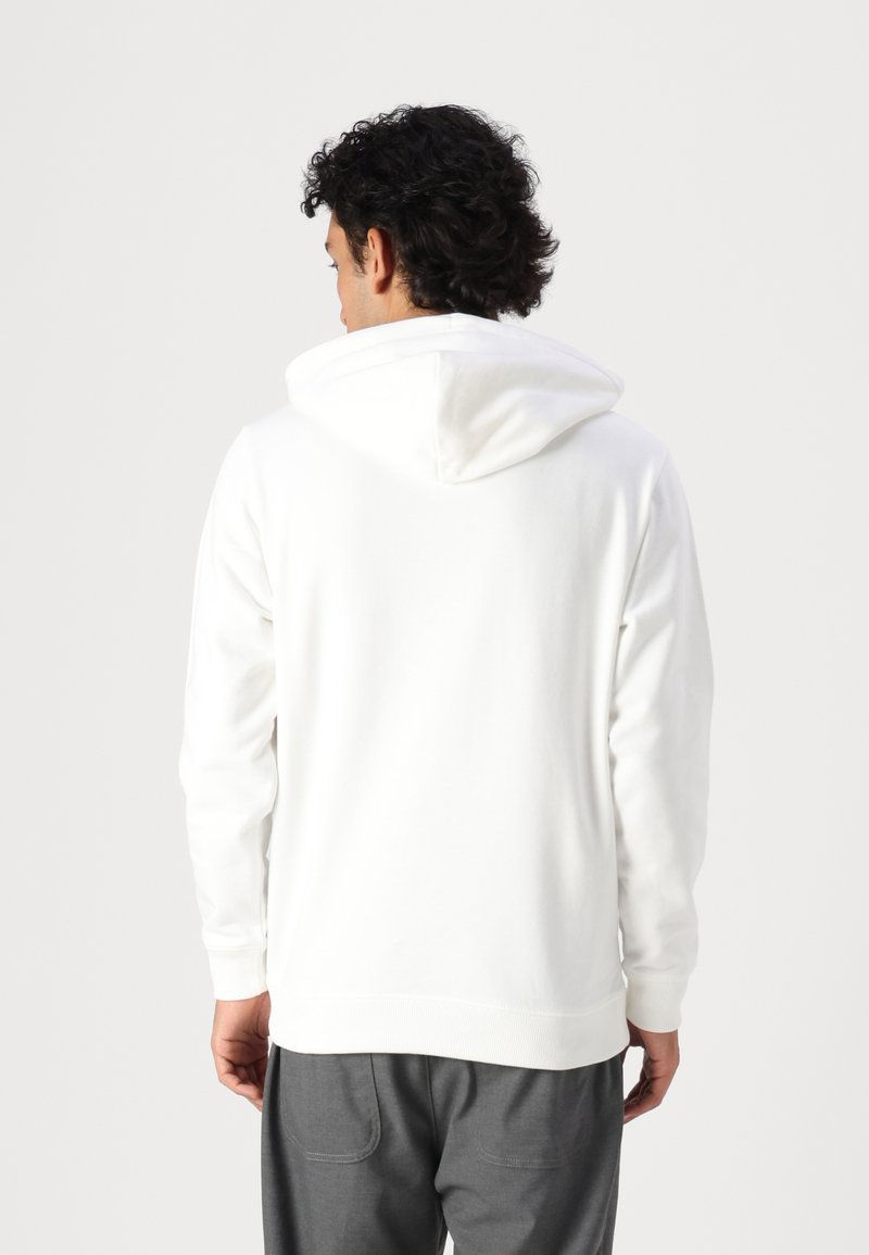Weißes Kapuzensweatshirt mit weicher Textur, ausgestattet mit einer Kängurutasche und gerippten Bündchen, gezeigt von hinten, kombiniert mit grauen Hosen.