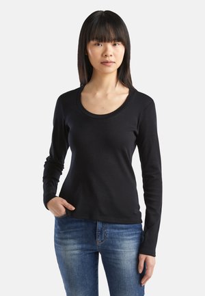 Schwarzes Langarmshirt mit rundem Ausschnitt, aus einem glatten Stoff gefertigt. Kombiniert mit blauen Jeans. Dekorative Nähte an den Handgelenken.