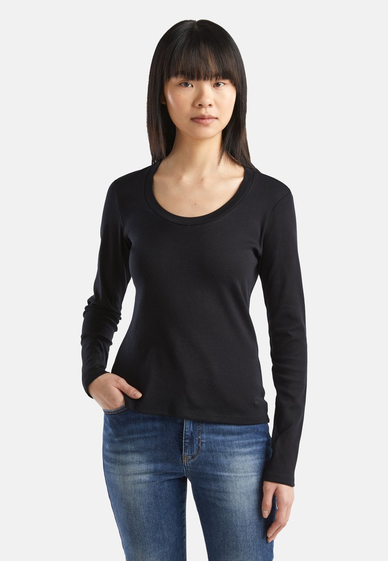 Schwarzes Langarmshirt mit rundem Ausschnitt, aus einem glatten Stoff gefertigt. Kombiniert mit blauen Jeans. Dekorative Nähte an den Handgelenken.