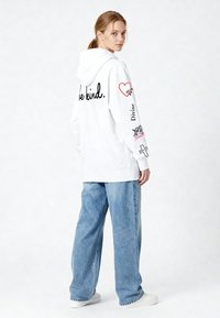 Bela majica s kapuco z napisom »bodi prijazen.« na hrbtni strani. Opremljena z grafičnimi potiski na rokavih. Kombinirana z ohlapnimi svetlo modrimi kavbojkami in belimi supergami.