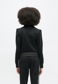 Μαύρο φούτερ με λείο ύφασμα, εφαρμοστή σιλουέτα και επένδυση στους ώμους. Διακρίνονται το ριμπ ζωνάκι στη μέση και τα μανίκια.