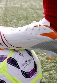 Biele futbalové kopačky s červenými a oranžovými akcentmi položené na viacfarebnej futbalovej lopte s fialovými a žltými detailmi. Viditeľný trávnik.