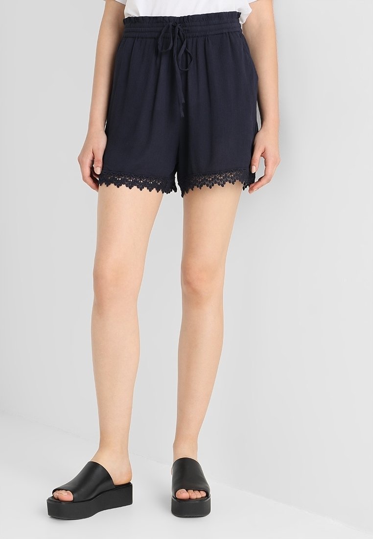 Shorts bleu marine en tissu léger, avec une coupe décontractée, une taille élastique et un bord en dentelle à l'ourlet. Portés avec des sandales noires à plateforme.