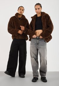 Twee mensen in bruine fuzzy jassen, de een met zwarte cargobroeken en de ander met grijze jeans en een zwarte crop top, staan zij aan zij.