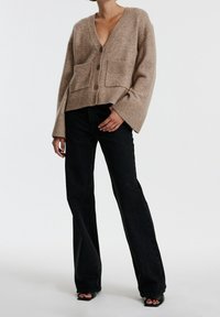 Cárdigan de punto beige con escote en V, dos bolsillos frontales y tres botones grandes. Combinado con unos vaqueros negros acampanados y tacones de punta abierta.