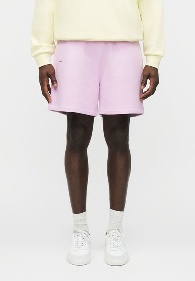 Mandlig model iført pastel lilla shorts, hvide sneakers, hvide sokker og en lysegul langærmet skjorte stående foran en hvid baggrund.
