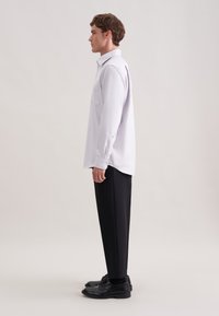 Camicia bianca a maniche lunghe con taschino e polsini abbottonati, abbinata a pantaloni neri su misura e scarpe nere, vista di lato.