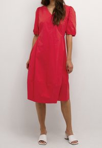 Nainen, jolla on polvipituinen punainen mekko, jossa on puffattuja lyhyitä hihoja, ja valkoiset avonaiset kärjet korkokenkä-mulerit, seisoo yksinkertaista taustaa vasten.