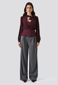 Blusa bordeaux a maniche lunghe con dettaglio a nodo e scollatura a chiave, abbinata a pantaloni grigi a vita alta e a palazzo plissettati.