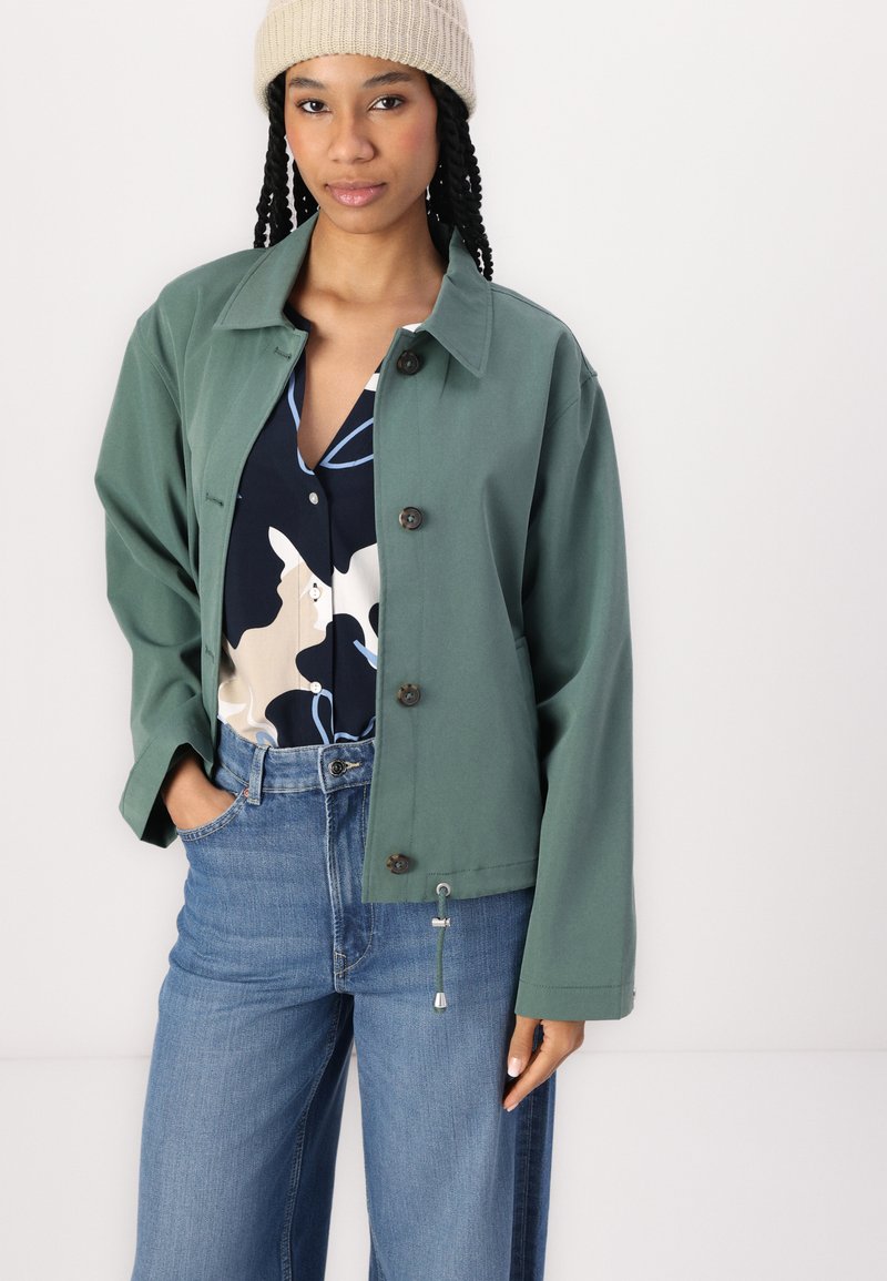 Groene, lichte jas met een kraag, knoopsluiting en koord aan de onderkant, gedragen over een gebloemde blouse en high-waisted jeans.