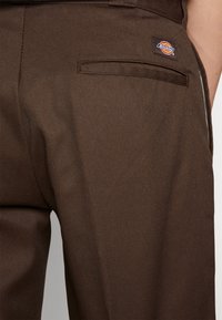 Brązowe spodnie materiałowe o gładkiej teksturze, z tylną kieszenią i małą, wielokolorową naszywką Dickies.