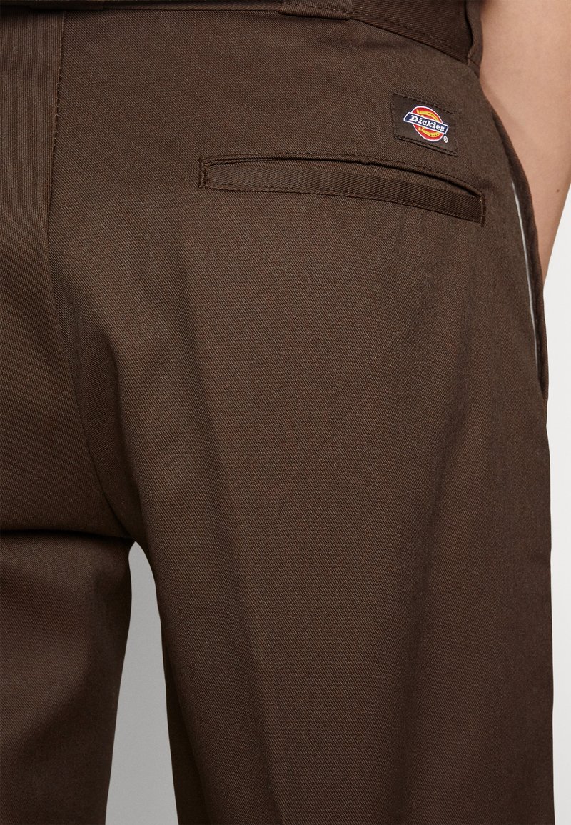 Pantalones de tela marrón con una textura suave, que cuentan con un bolsillo trasero y un pequeño parche de logo de Dickies multicolor.