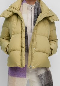 Jasnozielona kurtka puchowa z zapięciem na zamek, z dodatkowymi zatrzaskami oraz wysokim kołnierzem. Noszona na wielobarwnym szalu swetrowym.