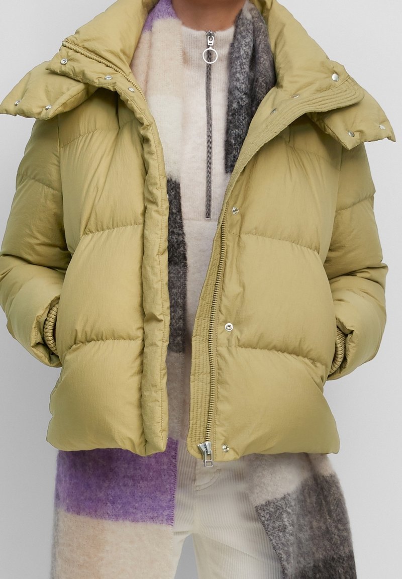 Jasnozielona kurtka puchowa z zapięciem na zamek, z dodatkowymi zatrzaskami oraz wysokim kołnierzem. Noszona na wielobarwnym szalu swetrowym.