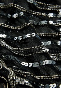 Close-up af sort netstof dekoreret med skinnende sølvglimmer og små metalliske perler arrangeret i bølgende linjer.