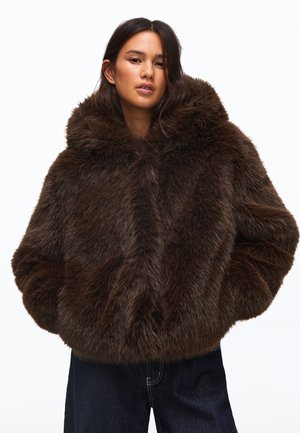 Femme portant une veste en fourrure marron foncé et duveteuse, les mains dans les poches et un pantalon foncé, debout devant un fond blanc.
