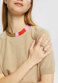 Suéter de punto beige con mangas cortas, que presenta un cuello acanalado multicolor. Tres anillos de plata llevados en los dedos. Textura suave.
