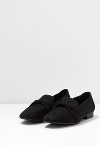 Ballerines en daim noir avec un bout pointu, présentant un design noué sur le dessus. Talon bas et texture lisse.