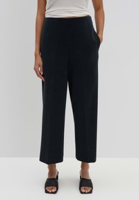 Pantalons larges en tissu bleu marine foncé, avec une taille haute, des poches latérales et une texture lisse. Associés à des sandales noires à bout ouvert.