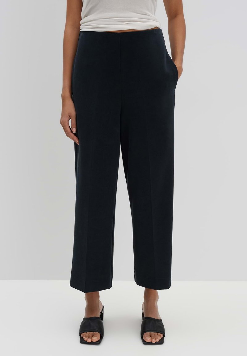Pantalons larges en tissu bleu marine foncé, avec une taille haute, des poches latérales et une texture lisse. Associés à des sandales noires à bout ouvert.