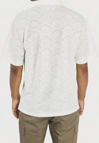 Biała koszula z krótkim rękawem z teksturowanym kwiatowym wzorem, z kołnierzykiem i luźnym krojem, połączona z oliwkowymi spodniami.