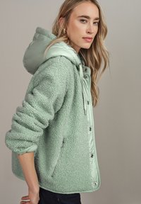 Donna con lunghi capelli biondi che indossa una giacca in pile verde chiaro con cappuccio e zip argentata, in piedi di lato contro uno sfondo neutro.