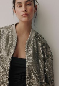 Sparkling silver bomber jacket with intricate sequin patterns; features a soft beige ribbed collar and textured fabric. Black top underneath.