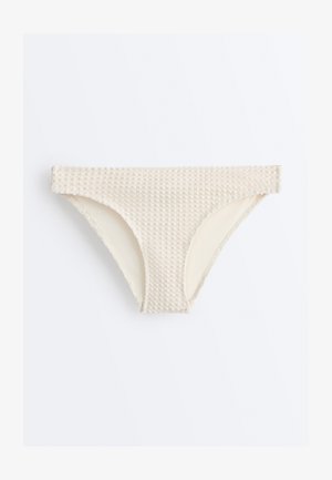 Bež bikini spodnji del z teksturo in z visokim izrezom noge, položen zravn na belo ozadje.