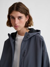 Jonge vrouw met kort bruin haar, met gouden oorringen, een wit overhemd en een donkergrijze ritsvest met capuchon.