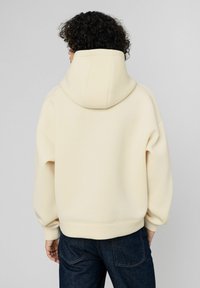 Sweat à capuche surdimensionné de couleur crème avec capuche à cordon, poignets et ourlet côtelés, fabriqué en tissu doux. Porté avec un jean bleu foncé.