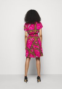 Růžové šaty s krátkými rukávy, s potiskem zlatých řetězů a geometrických tvarů, zúžené v pase, po kolena, v kombinaci s černými podpatky.