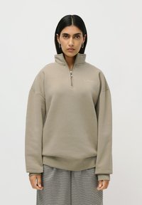 Kvinne med rett svart hår, iført en beige halv-zip genser og rutete vide bukser, som står mot en ensfarget hvit bakgrunn.