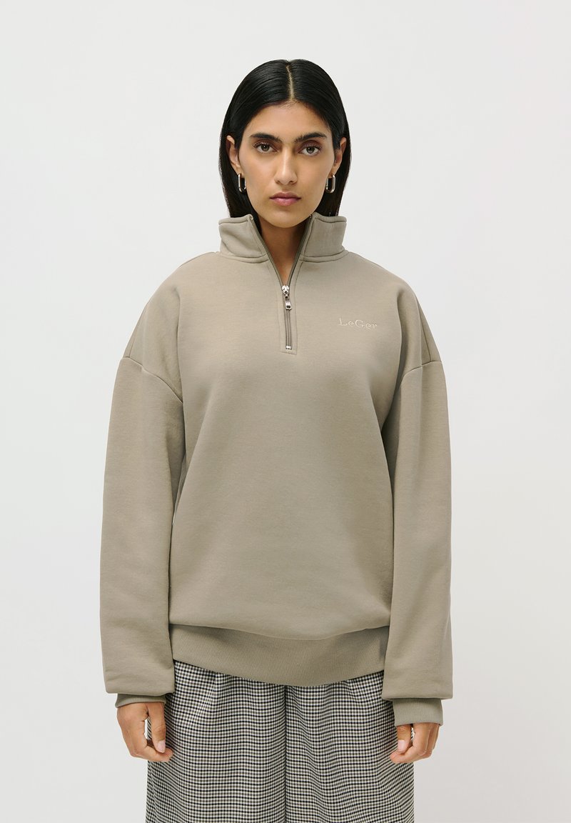 Kvinne med rett svart hår, iført en beige halv-zip genser og rutete vide bukser, som står mot en ensfarget hvit bakgrunn.