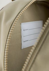 Beige väska med dragkedja som har ett inre med en vit etikett för namn och telefoninformation, med synliga sömmar längs kanterna.