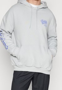 Hoodie grigio chiaro realizzato in tessuto morbido, con tasca frontale, cappuccio con coulisse e testo grafico blu sul petto e sulla manica.