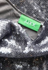 Szary materiał z cekinami z teksturowaną siateczkową nakładką. Posiada zieloną metkę z napisem "JJXX" w białym kolorze, przyszytą przy dekolcie.
