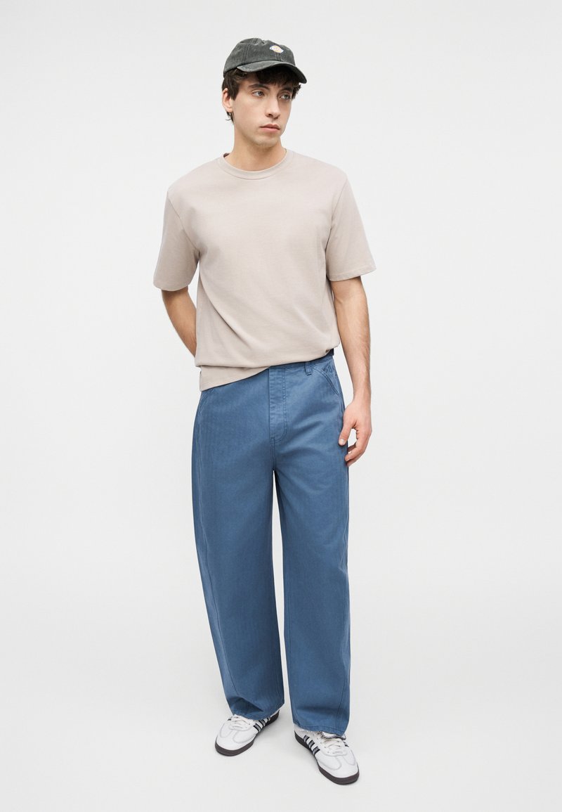 Young man wearing a beige t-shirt, loose blue pants, white sneakers, and a dark cap, standing against a plain white background.